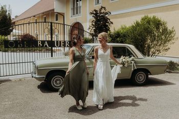 Braut und Begleiterin gehen lachend vor einem grünen Oldtimer entlang.